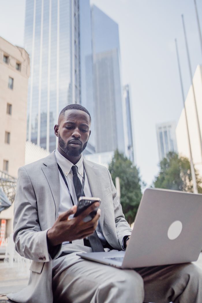 Young professional in a suit working with smartphone and laptop in urban setting.